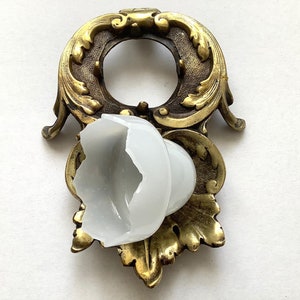 Könnte beinhalten: Ein goldfarbener, dekorativer Metallständer mit verzierten Details und einem weißen Glasschirm. Der Ständer hat eine kreisförmige Öffnung in der Mitte und ein florales Design.