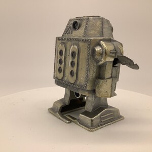 May include: A silver-colored toy robot with a wind-up key on the back. The robot has a rectangular body with a rounded head and a small, flat base.