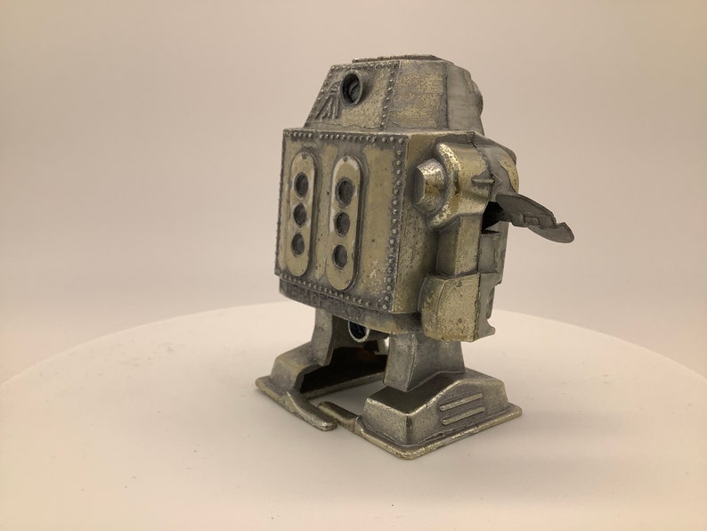 May include: A silver-colored toy robot with a wind-up key on the back. The robot has a rectangular body with a rounded head and a small, flat base.