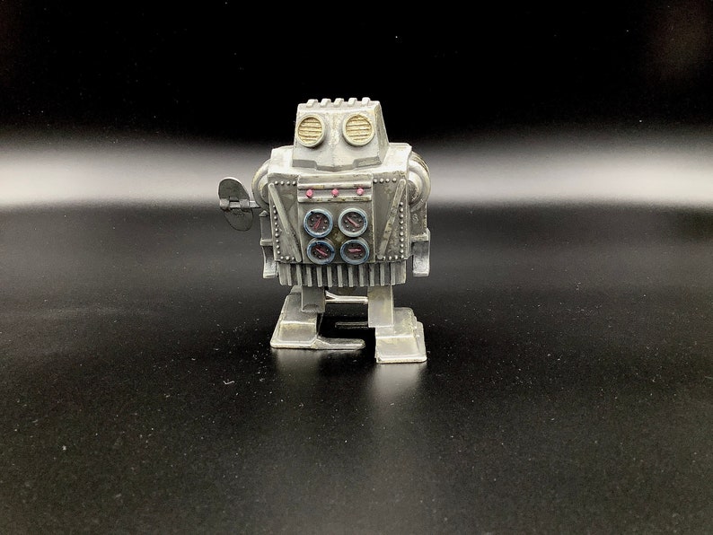 May include: A silver wind-up robot toy with a round head and four round eyes. The robot has a silver body with a black and white striped pattern and a silver base.