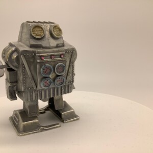 May include: A silver wind-up robot toy with a round head, two large eyes, and four gauges on its chest. The robot has a single arm with a round, flat hand.