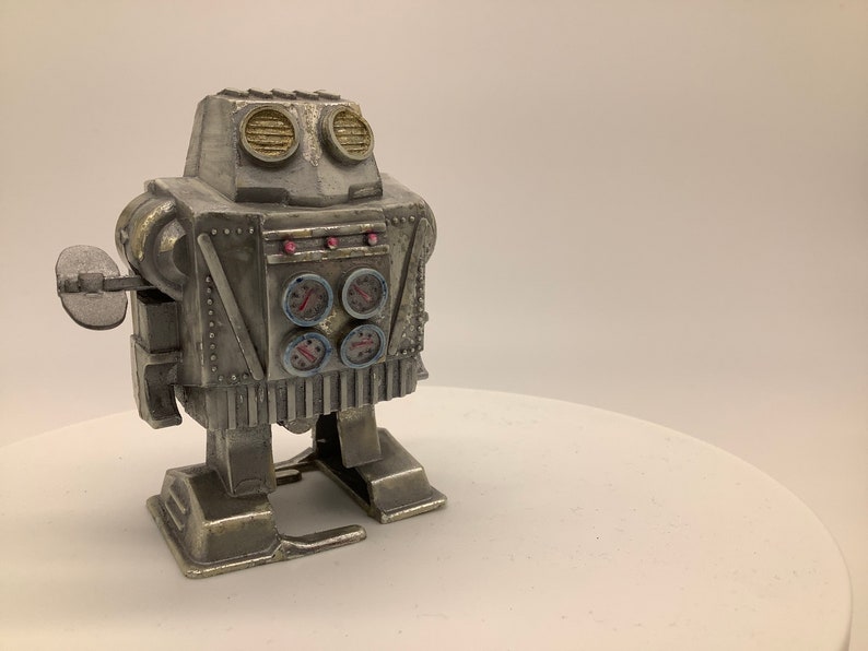 May include: A silver wind-up robot toy with a round head, two large eyes, and four gauges on its chest. The robot has a single arm with a round, flat hand.