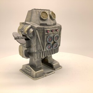 May include: A silver wind-up robot toy with a gold-colored head and arms. The robot has four round gauges on its chest and a propeller on its arm.
