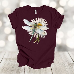 May include: A maroon t-shirt featuring a detailed illustration of a dragonfly resting on a white daisy. The dragonfly has a light blue body and wings, with green and black accents. The daisy has a yellow center.