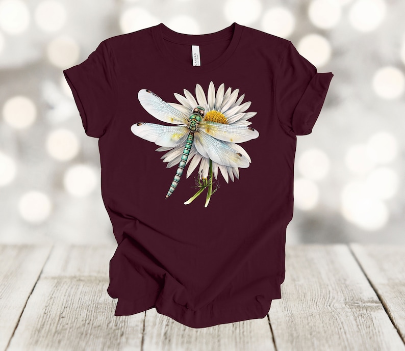 May include: A maroon t-shirt featuring a detailed illustration of a dragonfly resting on a white daisy. The dragonfly has a light blue body and wings, with green and black accents. The daisy has a yellow center.