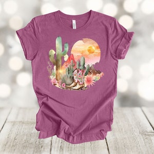 Camiseta gráfica Atardecer en el desierto: Camiseta estilo vaquero con estampado de cactus y botas. Heather Magenta