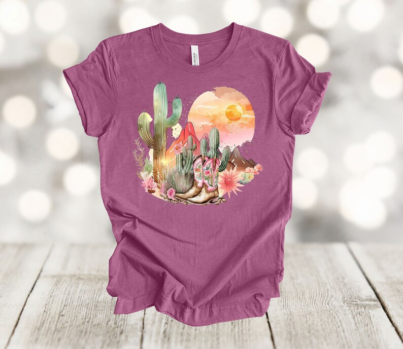 Camiseta gráfica Atardecer en el desierto: Camiseta estilo vaquero con estampado de cactus y botas. Heather Magenta