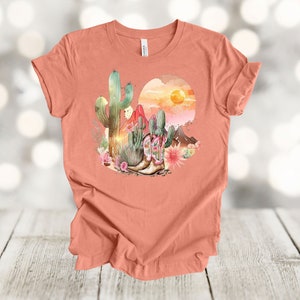 Camiseta gráfica Atardecer en el desierto: Camiseta estilo vaquero con estampado de cactus y botas. Heather Sunset