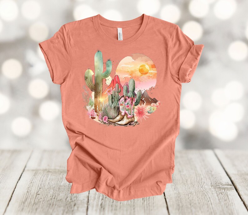 Camiseta gráfica Atardecer en el desierto: Camiseta estilo vaquero con estampado de cactus y botas. Heather Sunset
