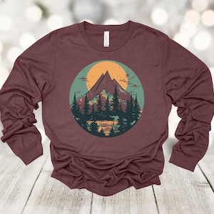 May include: A maroon long-sleeved shirt with a graphic print of a mountain range with a sunset behind it. The mountains are green and brown, and the sunset is orange and yellow. There are trees in front of the mountains.