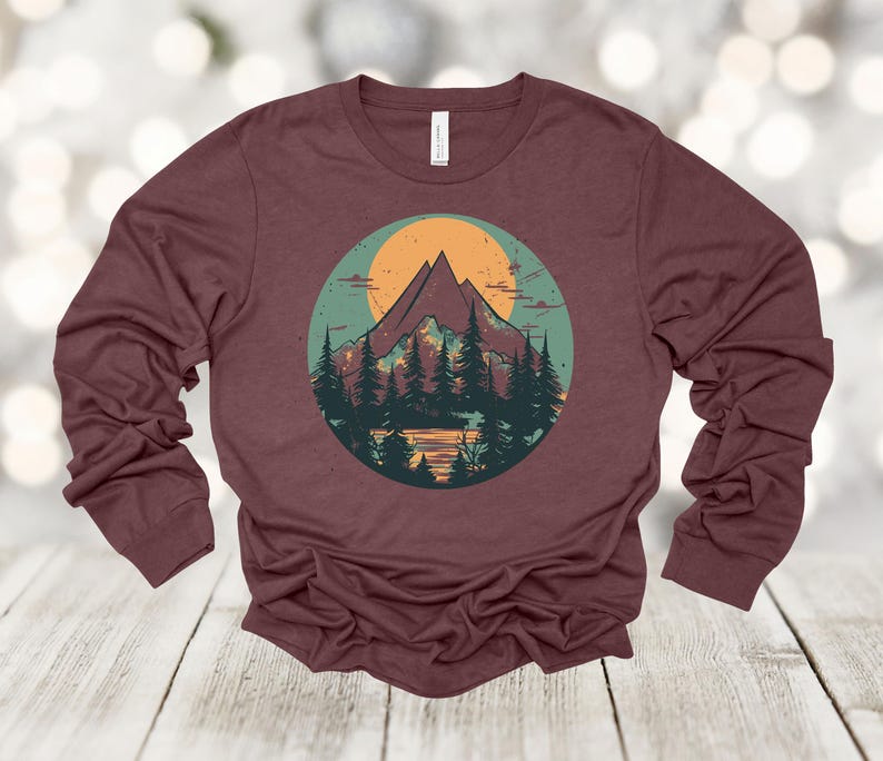 May include: A maroon long-sleeved shirt with a graphic print of a mountain range with a sunset behind it. The mountains are green and brown, and the sunset is orange and yellow. There are trees in front of the mountains.