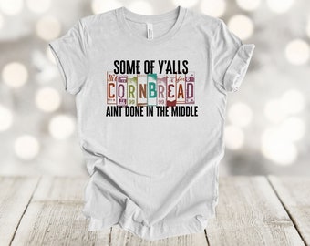May include: White t-shirt with the text "Some of y'alls cornbread ain't done in the middle" printed on it. The text is in a vintage style font and is surrounded by colorful squares.