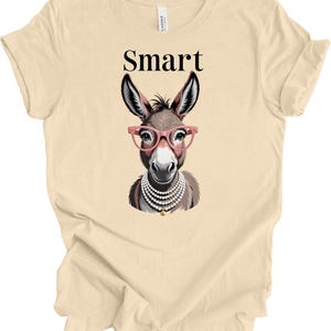 May include: Beige t-shirt featuring a cartoon donkey wearing pink glasses and a pearl necklace. The word "Smart" is printed above the donkey's head in black capital letters. The shirt is made of a soft, light-colored fabric.