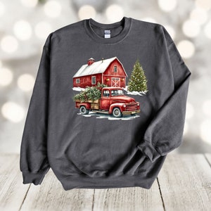 May include: A dark grey sweatshirt featuring a red truck carrying a Christmas tree in front of a red barn with a white roof. The truck is driving through a snowy scene.