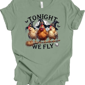 May include: Sage green t-shirt featuring a whimsical Halloween design. Three chickens wearing witch hats are perched on a broomstick, with the text "TONIGHT WE FLY" above and below the image. A full moon and bats complete the design.