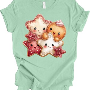 May include: A mint green t-shirt featuring a whimsical illustration of smiling gingerbread cookies and star-shaped cookies. The cookies have decorative icing and are in shades of red, white, and brown.