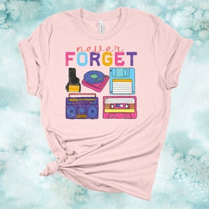 May include: Pink t-shirt with the text "never FORGET" in colourful letters. The shirt features a retro design with images of a cassette tape, a record player, a boombox, and a floppy disk.