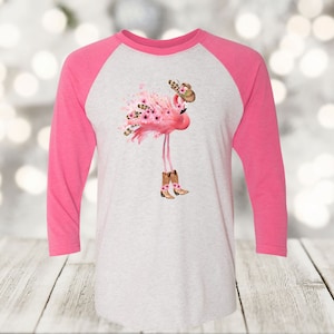 May include: A raglan-style baseball shirt with a pink and white design. The shirt features a pink flamingo wearing cowboy boots and a hat, adorned with flowers. The flamingo is the central graphic on the white torso of the shirt.