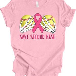 May include: Pink t-shirt featuring a graphic design with two yellow softballs held by white skeleton hands, a pink ribbon, and the text "SAVE SECOND BASE". The shirt is made of a soft material.