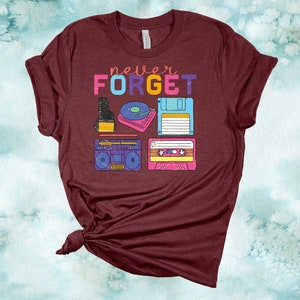 May include: A maroon t-shirt with the text "never FORGET" in rainbow colours. The shirt features a graphic of vintage music technology, including a cassette tape, a boombox, a floppy disk, a record player, and a camera.