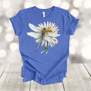 May include: A blue t-shirt featuring a detailed illustration of a dragonfly resting on a white daisy with a yellow center. The dragonfly has a green and blue body and delicate white wings. The shirt is displayed on a light wood surface.