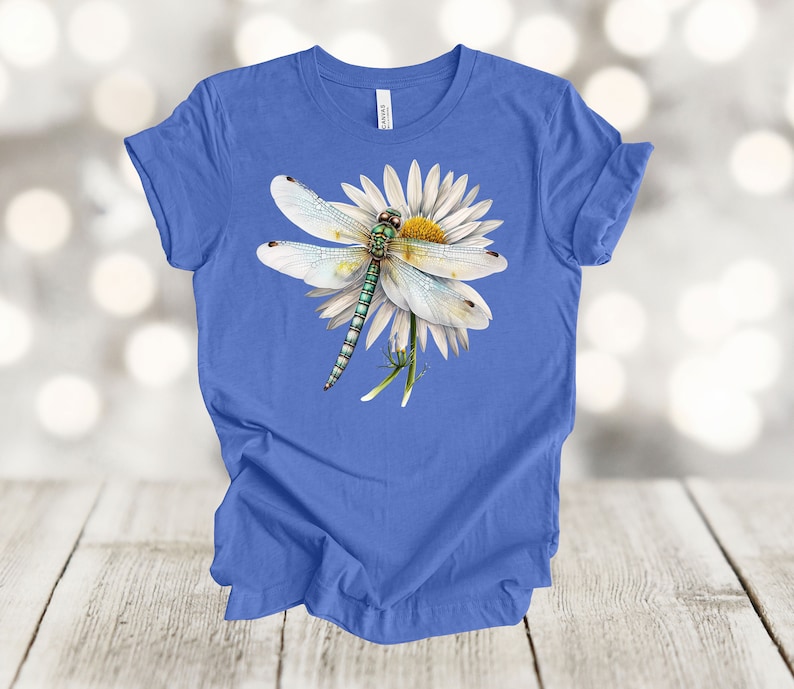 May include: A blue t-shirt featuring a detailed illustration of a dragonfly resting on a white daisy with a yellow center. The dragonfly has a green and blue body and delicate white wings. The shirt is displayed on a light wood surface.