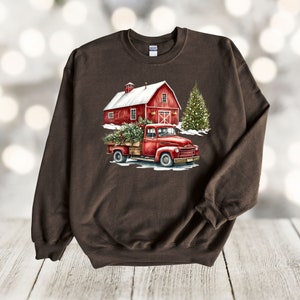 May include: A brown sweatshirt featuring a red truck carrying a Christmas tree in front of a red barn with a snowy roof. The truck is driving through a snowy scene with a Christmas tree in the background.