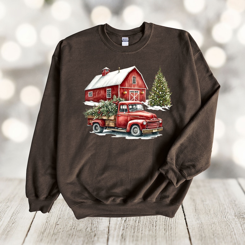 May include: A brown sweatshirt featuring a red truck carrying a Christmas tree in front of a red barn with a snowy roof. The truck is driving through a snowy scene with a Christmas tree in the background.