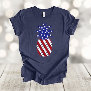 May include: Navy blue t-shirt featuring a patriotic pineapple design. The pineapple's crown is filled with white stars on a blue background, while the body has a red and white striped pattern. A casual tee for Independence Day.