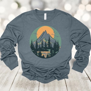 May include: A long-sleeved shirt with a blue background and a graphic of a mountain range with a yellow sun behind it. The mountains are green and brown, and the sun is yellow with orange rays.