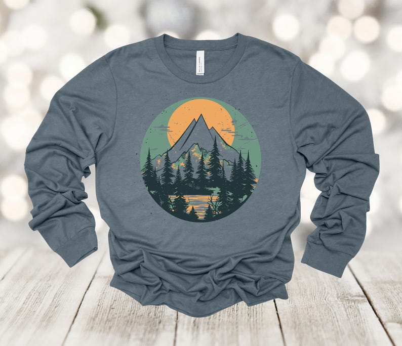 May include: A long-sleeved shirt with a blue background and a graphic of a mountain range with a yellow sun behind it. The mountains are green and brown, and the sun is yellow with orange rays.