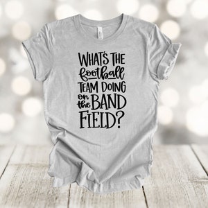 May include: A gray t-shirt with black text that reads "What's the football team doing on the band field?"