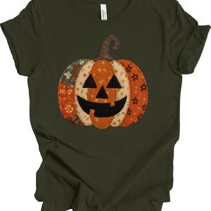May include: Dark green t-shirt featuring a patchwork pumpkin design. The pumpkin has a smiling jack-o'-lantern face with black eyes and a mouth. The pumpkin is orange, cream, and brown with floral and patterned fabric accents. The shirt is made of a soft material.