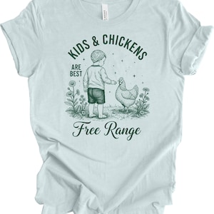 May include: Light blue t-shirt with a graphic featuring a child and a chicken, surrounded by flowers. The text reads "KIDS & CHICKENS ARE BEST" and "Free Range". The design is in a vintage style.