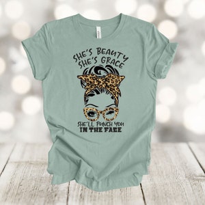 May include: A light green t-shirt with a graphic of a woman with a leopard print headband and sunglasses. The text on the shirt reads "She's beauty she's grace she'll punch you in the face."