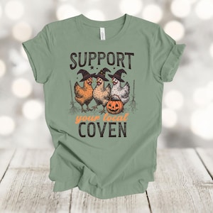 May include: Sage green t-shirt featuring a graphic of three chickens wearing witch hats, a carved pumpkin, and the text "Support your local Coven." The design is in black and orange.