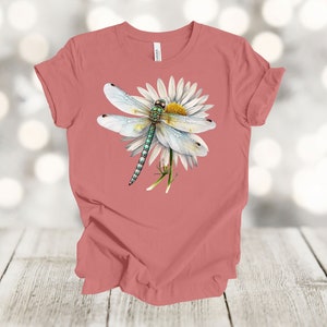 May include: A coral-colored t-shirt featuring a detailed illustration of a dragonfly perched on a white daisy. The dragonfly has delicate, translucent wings and a green and blue body. The daisy has a yellow center.