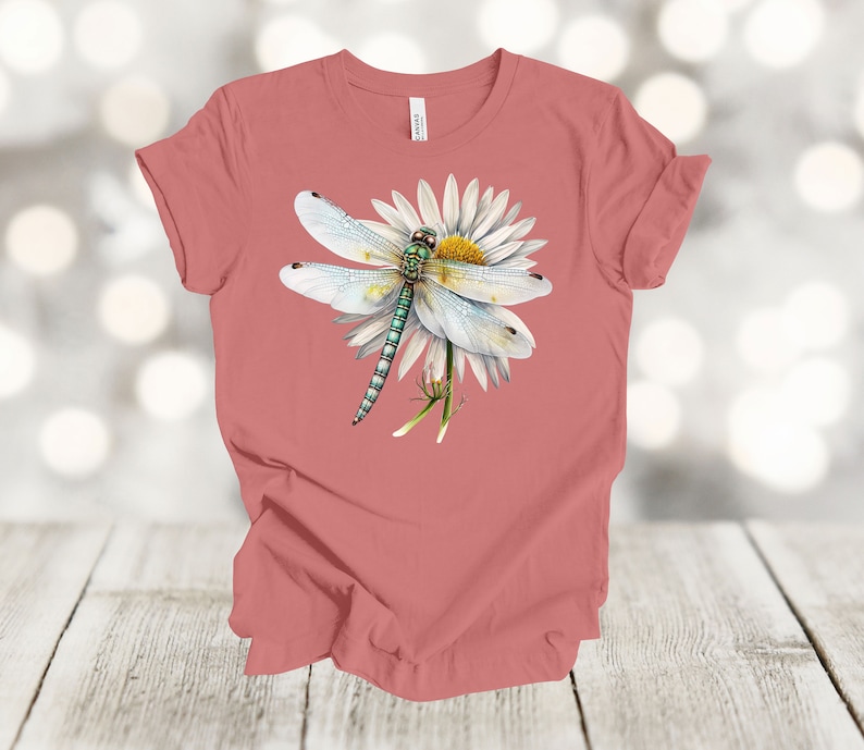 May include: A coral-colored t-shirt featuring a detailed illustration of a dragonfly perched on a white daisy. The dragonfly has delicate, translucent wings and a green and blue body. The daisy has a yellow center.