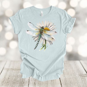 May include: Light blue t-shirt featuring a detailed illustration of a dragonfly resting on a white daisy. The dragonfly has a green body and translucent wings. The daisy has a yellow center and white petals. The shirt is made of a soft material.
