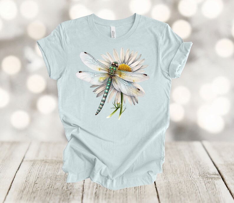 May include: Light blue t-shirt featuring a detailed illustration of a dragonfly resting on a white daisy. The dragonfly has a green body and translucent wings. The daisy has a yellow center and white petals. The shirt is made of a soft material.