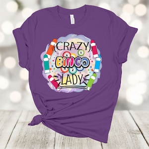 May include: Purple t-shirt with a colorful graphic design that says "(Crazy) Bingo Lady" with bingo balls and stars.