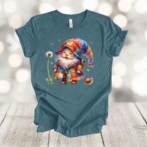 May include: A teal blue t-shirt with a colorful gnome wearing a patchwork hat and carrying a backpack. The gnome is smiling and standing next to a dandelion and a snail.