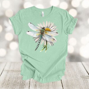 May include: A light green t-shirt featuring a detailed illustration of a dragonfly perched on a white daisy with a yellow center. The dragonfly has white wings and a green and blue body. The shirt is made of soft material.