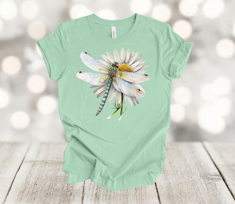 May include: A light green t-shirt featuring a detailed illustration of a dragonfly perched on a white daisy with a yellow center. The dragonfly has white wings and a green and blue body. The shirt is made of soft material.