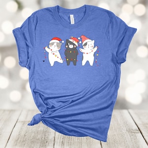 May include: A blue t-shirt with a graphic of three cats wearing Santa hats and holding up Christmas lights.