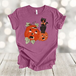 May include: A purple t-shirt with a black cat wearing a bow tie sitting on top of two orange jack-o'-lantern pumpkins.