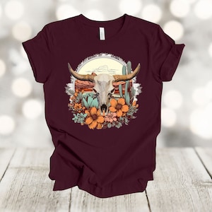 May include: A maroon t-shirt with a graphic of a white bull skull surrounded by orange and green flowers and cacti. The graphic is set against a sunset background.