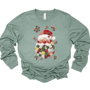 May include: A long-sleeved, sage green shirt featuring a festive Santa Claus design. The Santa Claus graphic is made of patchwork fabric with red, green, and white patterns. The Santa Claus wears a red hat with a white trim and a star.