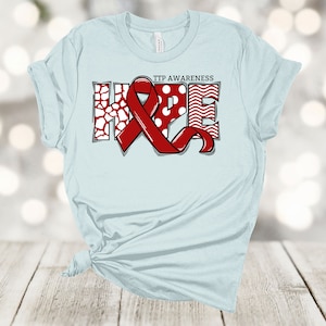 May include: Light blue t-shirt with the word "HOPE" in red, white, and black patterned letters. A red ribbon is tied around the letters. The text "TTP AWARENESS" is printed above the word "HOPE".