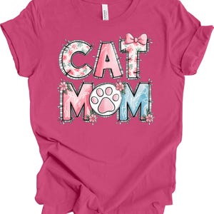 May include: A pink t-shirt with the words "CAT MOM" in a colorful, floral design. The letters are outlined with a stitched effect, and a cat paw print is in the center. A pink bow is above the letter "T".